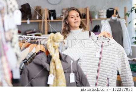 Stylish young girl standing in clothing shop and looking new down jacket made of high-quality material. Shopping and seasonal sales concept, winter season Stylish young girl standing in clothing shop and looking new down jacket made of high-quality material. Shopping and seasonal sales concept, winter season 132257955