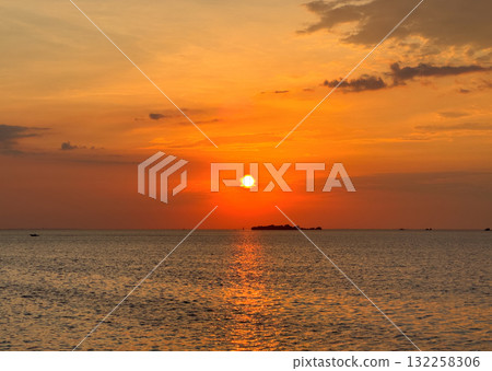 The sun sets over the sea, casting a warm glow across the water and sky. The scene is serene and picturesque, with clouds reflecting the golden hues of the sunset. 132258306