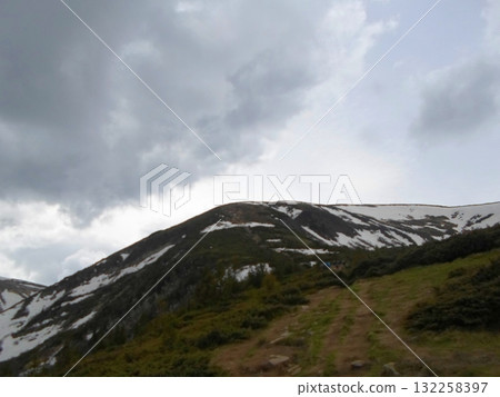 Snowy Mountain Trail with Patches of Greenery Under a Cloudy Sky 132258397