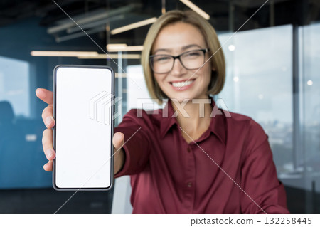 Smiling businesswoman in office presenting a smartphone with a blank white screen, perfect mockup for mobile apps, services or digital content with space for copy and branding 132258445