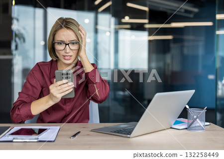 Businesswoman looking distressed and clutching her head while reading upsetting information on a smartphone, experiencing work stress and anxiety in a modern office environment Businesswoman looking distressed and clutching her head while reading upsetting information on a smartphone, experiencing work stress and anxiety in a modern office environment 132258449