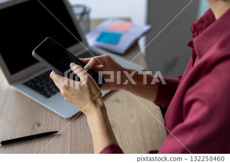 Woman's hands engaging with a smartphone while working at a desk, with a laptop open in the background, illustrating communication and business technology 132258460