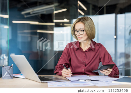 Businesswoman calculating finances using a calculator, signing documents and managing paperwork at a desk while working in a contemporary office environment Businesswoman calculating finances using a calculator, signing documents and managing paperwork at a desk while working in a contemporary office environment 132258496