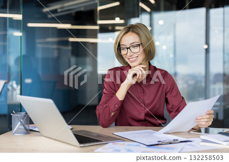 Young asian businesswoman in glasses smiling happily while reviewing documents and working with a laptop at her desk in a contemporary corporate office 132258503