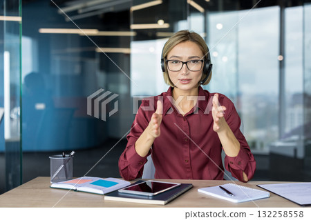 Woman wearing a headset and glasses sitting at a desk, looking at the camera and gesturing with her hands while engaging in an online business meeting, video call, or webinar 132258558