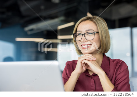 Happy professional businesswoman wearing eyeglasses and a maroon shirt, smiling cheerfully while working on a laptop in a modern corporate office environment 132258564