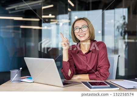 Happy businesswoman wearing glasses working on laptop in modern office, having a sudden idea and pointing finger up, feeling inspiration and eureka moment 132258574