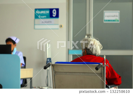 An elderly woman with gray hair in a red dress sits in a wheelchair and waits in a hospital examination room with a sign in Thai. A nurse stands behind her. An elderly woman with gray hair in a red dress sits in a wheelchair and waits in a hospital examination room with a sign in Thai. A nurse stands behind her. 132258610