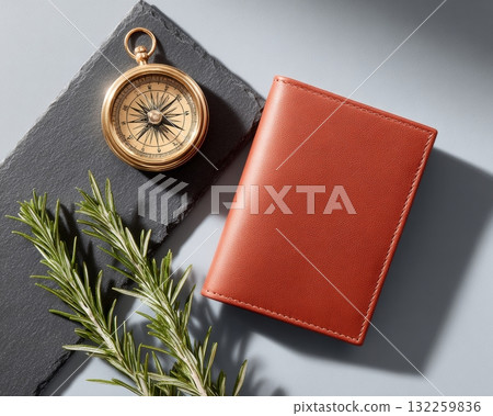 Top-down view of burnt sienna mycelium-based leather wallet with brass compass and rosemary on slate in editorial flat lay 132259836