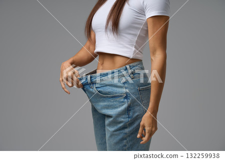 Close-up of a fit young woman in a white crop top pulling out the waistband of loose jeans, symbolizing successful weight loss. Studio shot with neutral gray background. 132259938