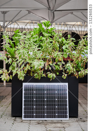Photovoltaic solar panel at large planter filled with vibrant green foliage, set on paved area under canopy. Urban setting emphasizes integration of green energy solutions within modern cityscapes. Photovoltaic solar panel at large planter filled with vibrant green foliage, set on paved area under canopy. Urban setting emphasizes integration of green energy solutions within modern cityscapes. 132260833