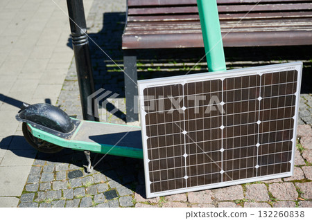 Mint-green electric scooter rests on cobblestone path, with solar panel leaning against it. Solar panel strategically placed to capture sunlight, highlighting sustainable method to charge scooter. 132260838