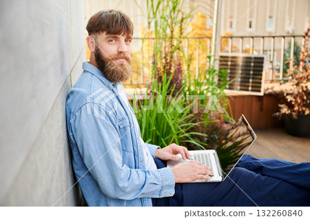 Portrait of happy bearded man works on laptop on lap. Solar panel nearby, emphasizing sustainable, modern eco-friendly workspace that combines technology and sustainability. 132260840