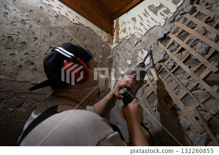 A construction worker in protective gear uses a hammer drill to remove old plaster and cement from an interior wall. A man uses a hammer drill to remove old tiles and cement from the walls. A construction worker in protective gear uses a hammer drill to remove old plaster and cement from an interior wall. A man uses a hammer drill to remove old tiles and cement from the walls. 132260855