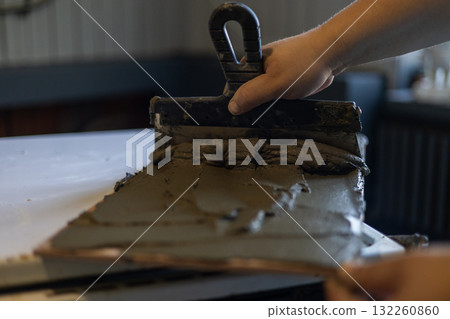 A worker manually applies cement-based tile adhesive with a notched trowel, evenly distributing the mortar over ceramic or porcelain tiles during a home renovation. A worker manually applies cement-based tile adhesive with a notched trowel, evenly distributing the mortar over ceramic or porcelain tiles during a home renovation. 132260860