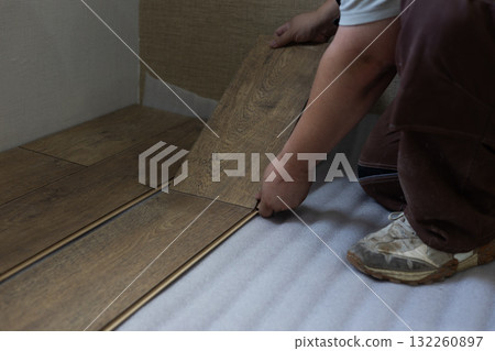 A man installs new wood-look laminate flooring, carefully joining the planks onto a white backing during a home renovation or construction project. A man lays laminate flooring on a house floor. 132260897