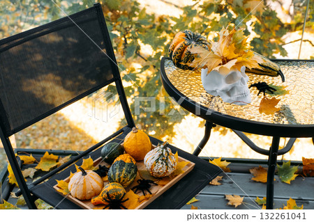 Decorative gourds, plastic spiders, and a white skull with autumn leaves set on a small balcony table and chair. Halloween home styling cozy seasonal balcony, creative fall decor, outdoor mood setting 132261041