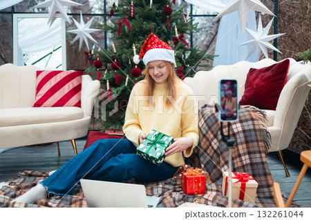 Smiling blonde woman in Santa hat showing Christmas gift during video call 132261044