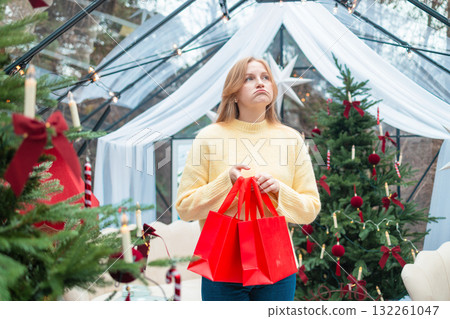 Thoughtful blonde woman holding red shopping bags during Christmas holidays Thoughtful blonde woman holding red shopping bags during Christmas holidays 132261047