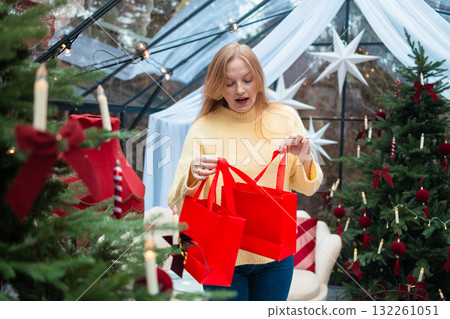Surprised blonde woman looking into red shopping bags near Christmas tree 132261051