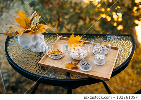 Outdoor glass table setup featuring a skull adorned with autumn leaves next to a wooden tray filled with assorted supplements. Mortality and health awareness in autumn visuals, memento mori, 132261057