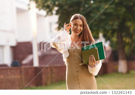 With notepads. Beautiful female student is outdoors near the university With notepads. Beautiful female student is outdoors near the university 132261143