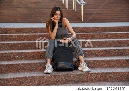 Holding smartphone, sitting on the stairs. Beautiful female student is outdoors near the university Holding smartphone, sitting on the stairs. Beautiful female student is outdoors near the university 132261161