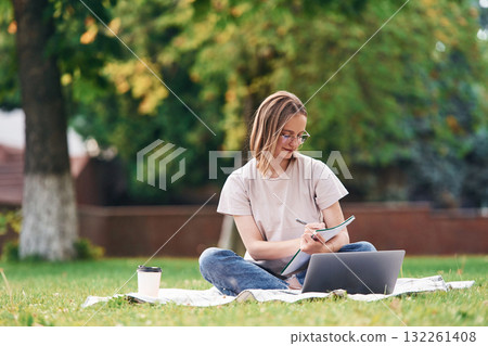 On the grass with laptop. Beautiful female student is outdoors near the university On the grass with laptop. Beautiful female student is outdoors near the university 132261408