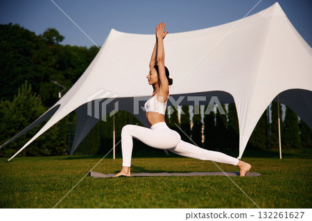 White tent behind, doing yoga. Attractive young fitness woman is on the green grass at daytime 132261627