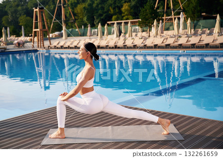 Busy by doing yoga exercise. Fitness woman is near the pool Busy by doing yoga exercise. Fitness woman is near the pool 132261659