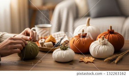 Hands sewing a handmade fabric pumpkin for autumn decor on a wooden table with craft supplies and a hot drink. 132263162