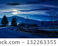 country road in mountains at night. beautiful countryside landscape in late autumn. fir trees on the roadside in full moon light. scenic view of blue sky with clouds above the distant ridge in snow 132263355