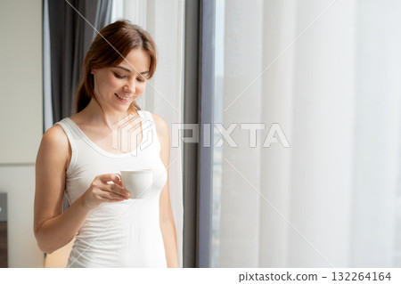 Happy young caucasian woman standing drinking coffee in morning in bedroom at home. 132264164