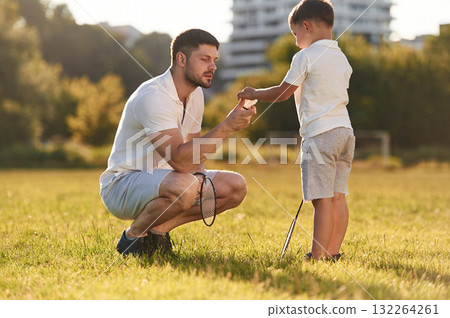 With badminton rackets. Father and little son are together on the summer field With badminton rackets. Father and little son are together on the summer field 132264261