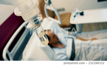 Intravenous drip tube with a blurred patient lying in hospital bed in the background. Medical care, recovery, health treatment, hospital room scene, IV therapy concept Intravenous drip tube with a blurred patient lying in hospital bed in the background. Medical care, recovery, health treatment, hospital room scene, IV therapy concept 132266332