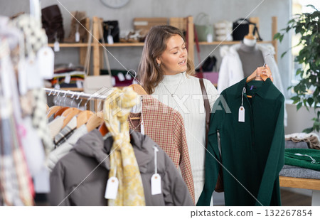 Positive young girl standing in interior of clothing boutique and choosing dress for autumn-winter season Positive young girl standing in interior of clothing boutique and choosing dress for autumn-winter season 132267854