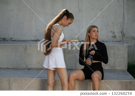 Holding smartphone, taking a break. Two sportive women are outdoors together 132268259