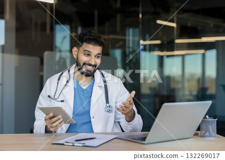 Male doctor in a lab coat with stethoscope conducts a telehealth video consultation from an office, using laptop and tablet to discuss patient care and medical advice remotely 132269017