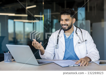Indian male doctor in white coat with stethoscope smiling and gesturing at laptop during a virtual video consultation, offering telemedicine advice and remote healthcare support Indian male doctor in white coat with stethoscope smiling and gesturing at laptop during a virtual video consultation, offering telemedicine advice and remote healthcare support 132269023