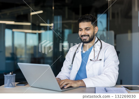 Male indian doctor smiling at laptop while providing telehealth consultation from office desk, confident and professional during virtual patient care and remote medical support 132269030