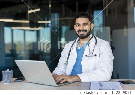 Young indian male physician wearing a white coat and stethoscope, smiling while working on a laptop at an office desk, symbolizing modern healthcare, telehealth, and accessible medical consultations 132269031