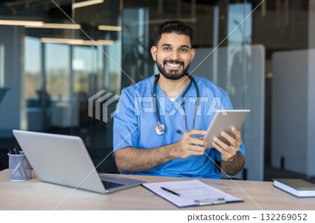 Young indian male doctor in blue scrubs and a stethoscope smiling. Holding a digital tablet while sitting at a desk with a laptop. Representing modern healthcare technology and online medical services 132269052