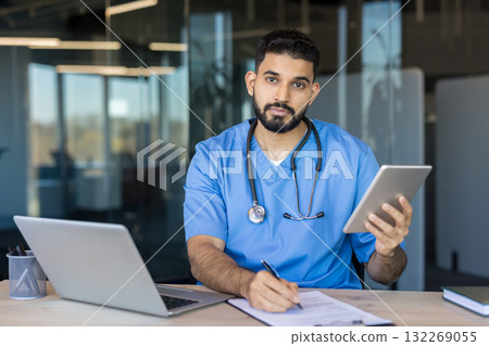 Male doctor in blue scrubs and stethoscope sits at desk with tablet and laptop, writing notes-focused portrait conveying modern telemedicine, digital documentation, and clinical care 132269055