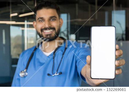 Smiling male doctor wearing blue scrubs and stethoscope holding up a smartphone with a blank white screen, symbolizing telecommunication, smart technology, and healthcare applications 132269081