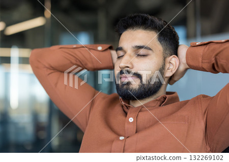 Young bearded man in a brown shirt relaxing in an office, resting with closed eyes and hands behind his head, finding peace and stress relief during a hard workday 132269102