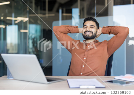 Happy young bearded businessman enjoying a break and relaxing at his office desk with eyes closed, smiling, showing satisfaction after completing work 132269103