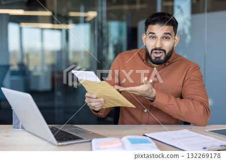 Young businessman sitting at desk in office receiving surprising news while opening an important document from a brown envelope, reacting with a shocked expression 132269178
