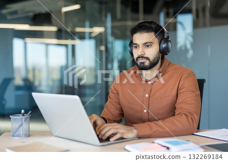 Young indian businessman wearing headphones, focusing intently while typing on a laptop computer, working at a clean desk in a contemporary open-plan office environment Young indian businessman wearing headphones, focusing intently while typing on a laptop computer, working at a clean desk in a contemporary open-plan office environment 132269184