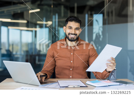Smiling indian businessman sitting at office desk, reviewing financial documents and using a laptop, representing success, productivity, confidence, and modern business Smiling indian businessman sitting at office desk, reviewing financial documents and using a laptop, representing success, productivity, confidence, and modern business 132269225