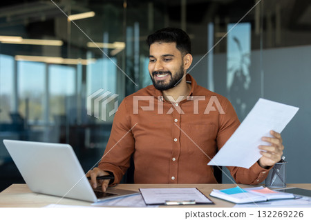 Smiling business professional reviewing documents while working on a laptop in a contemporary office environment, conveying productivity and professionalism. Smiling business professional reviewing documents while working on a laptop in a contemporary office environment, conveying productivity and professionalism. 132269226